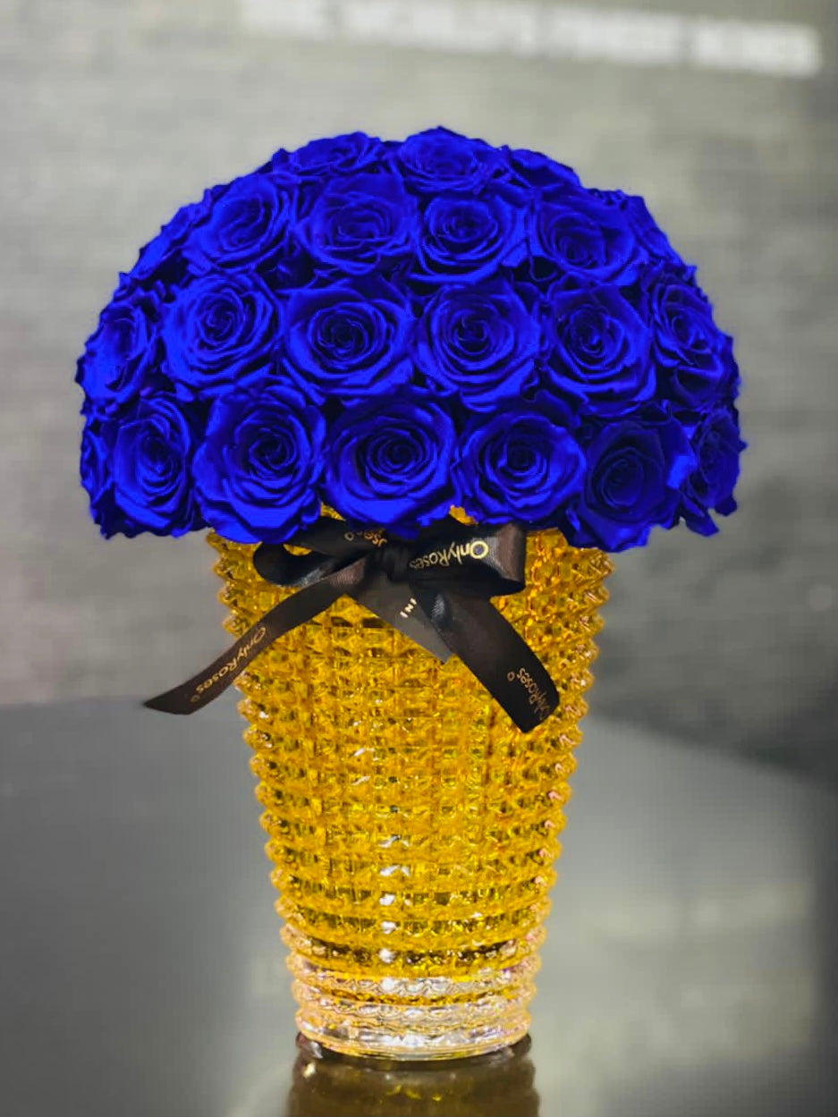 Bouquet of blue roses in a gold vase on a reflective surface with a blurred background