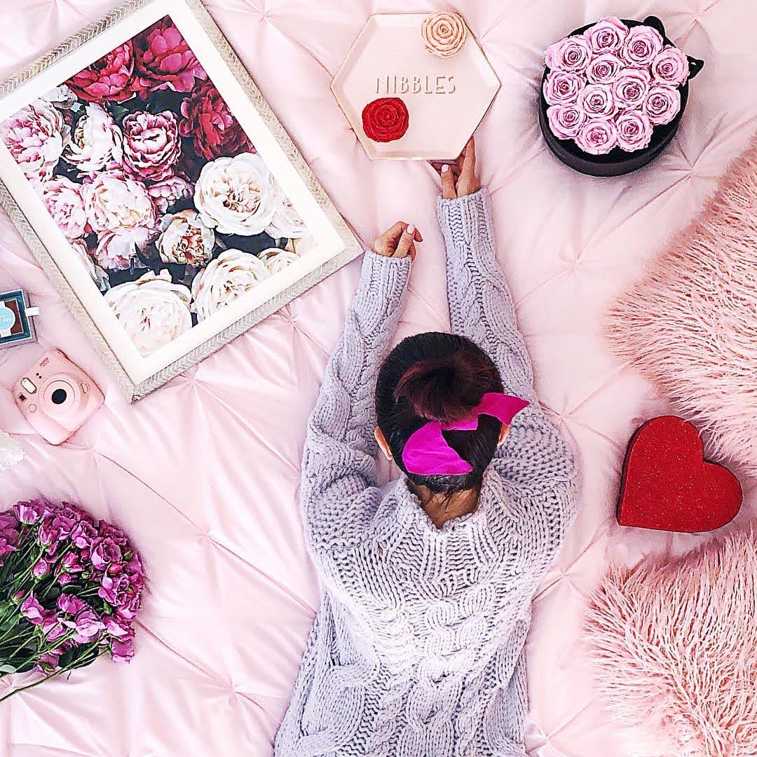 Person lying on a pink surface with flowers, a heart-shaped box, and a framed floral picture.