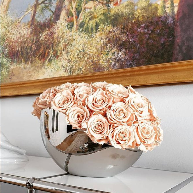 Bouquet of peach roses in a silver bowl on a white surface with a painting in the background