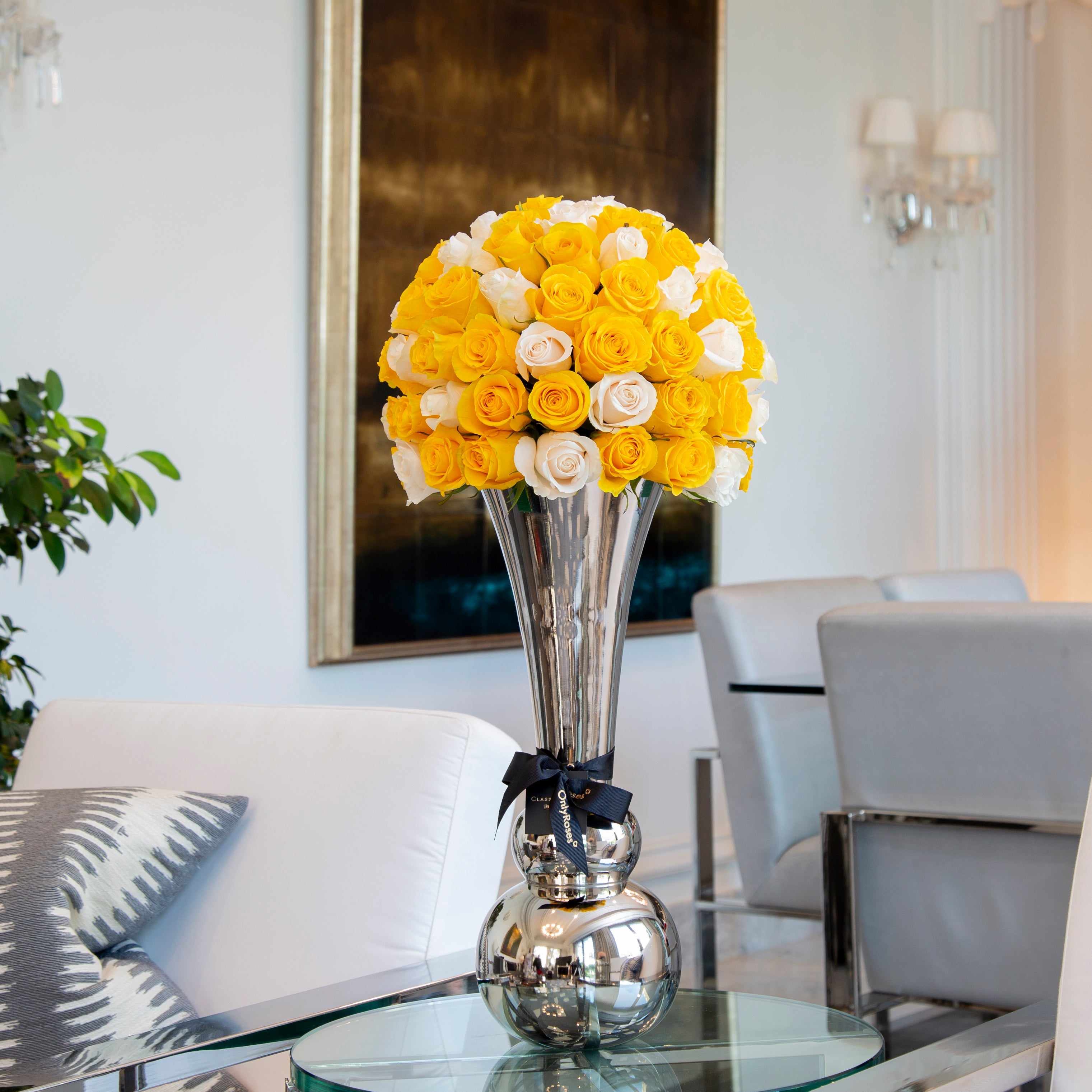 Bouquet of yellow and white roses in a silver vase on a glass table in a modern living room.