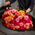 Heart-shaped arrangement of pink, orange, and red roses on a dark surface with a blurred background.