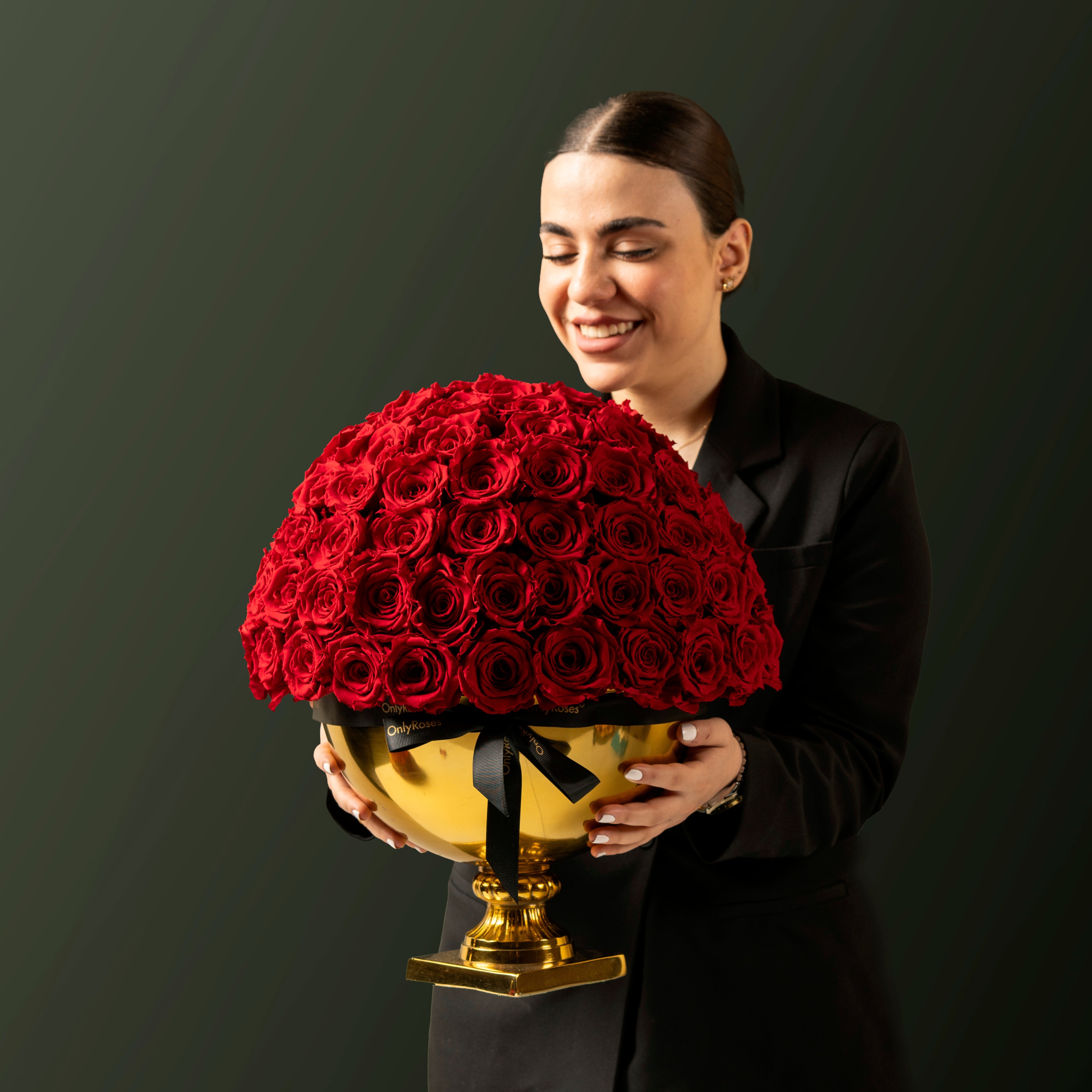 Woman holding a large bouquet of red roses in a gold vase against a dark background