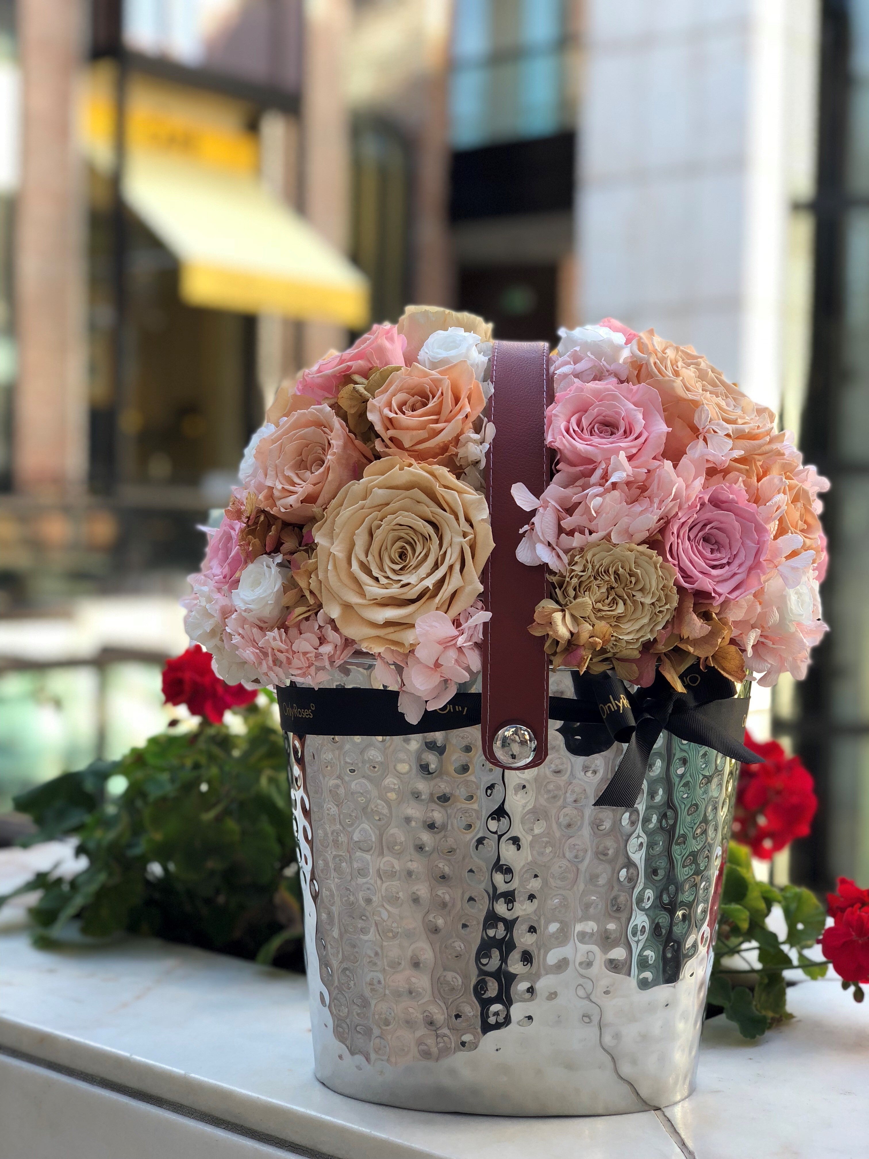 Decorative flower arrangement in a metal bucket with a brown ribbon against a blurred urban background.