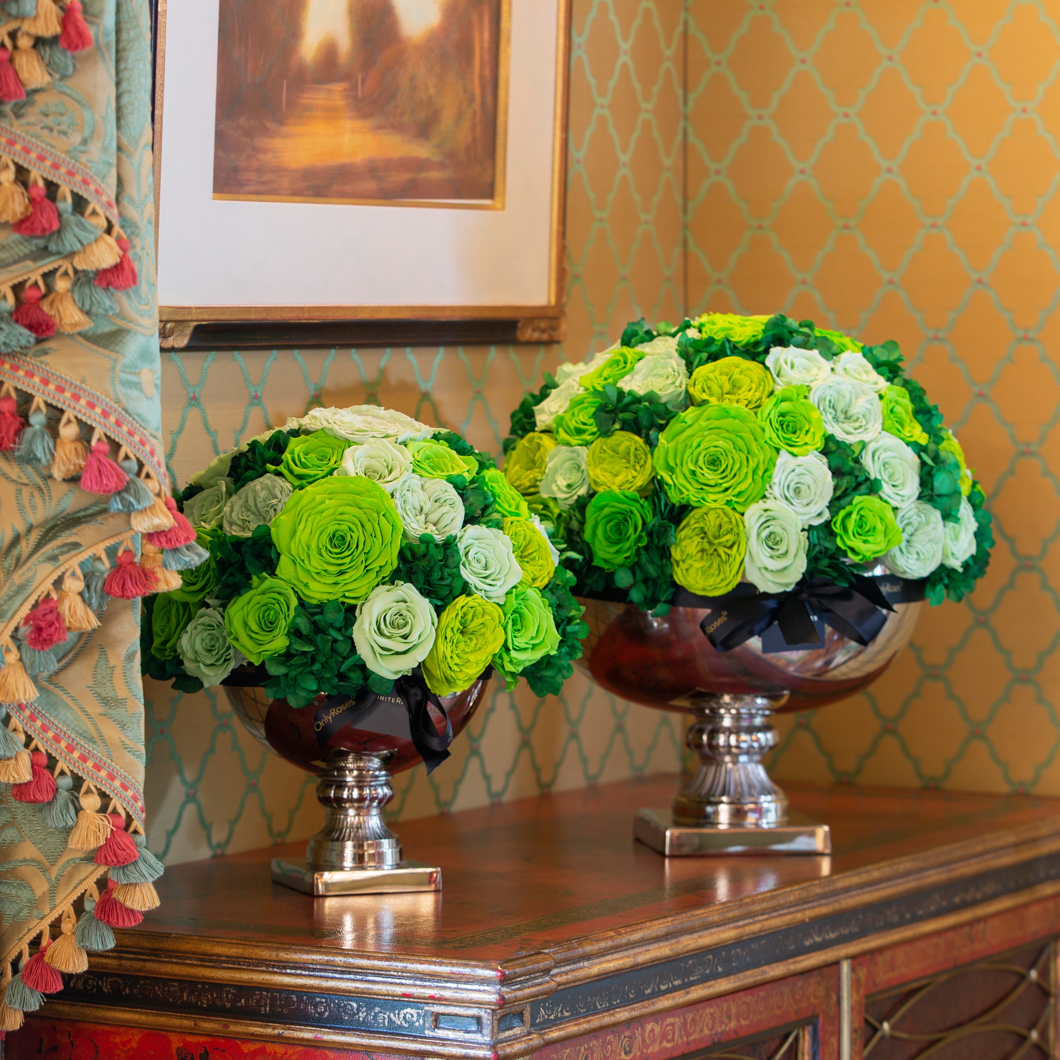 Two floral arrangements in silver urns on a decorative table with patterned wallpaper.