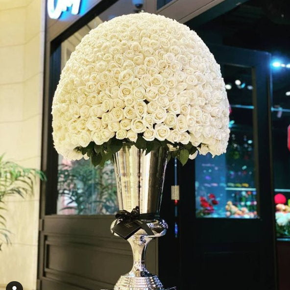 Large decorative arrangement of white flowers in a silver vase against a blurred indoor background.