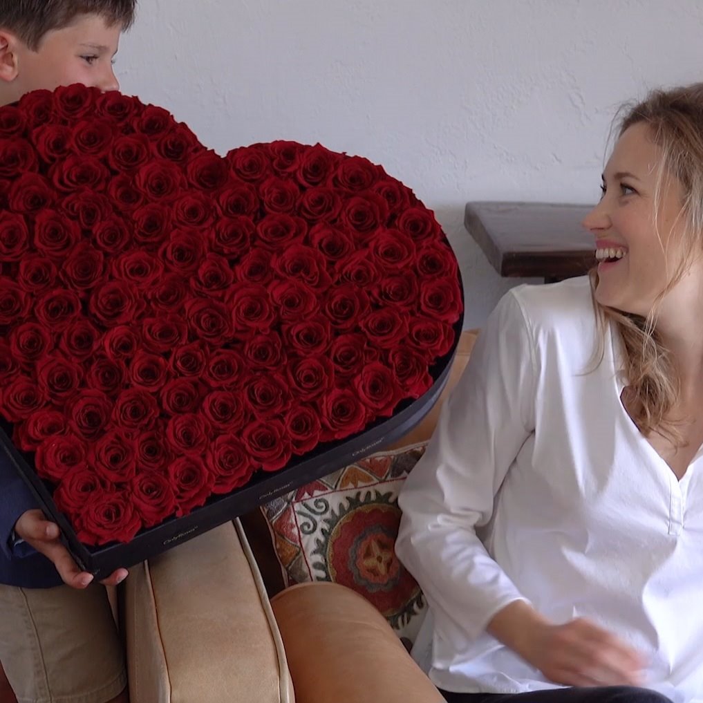 Person holding a heart-shaped box of red roses to a smiling woman.