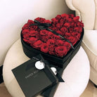 Heart-shaped box of red roses with a black box on a white surface.