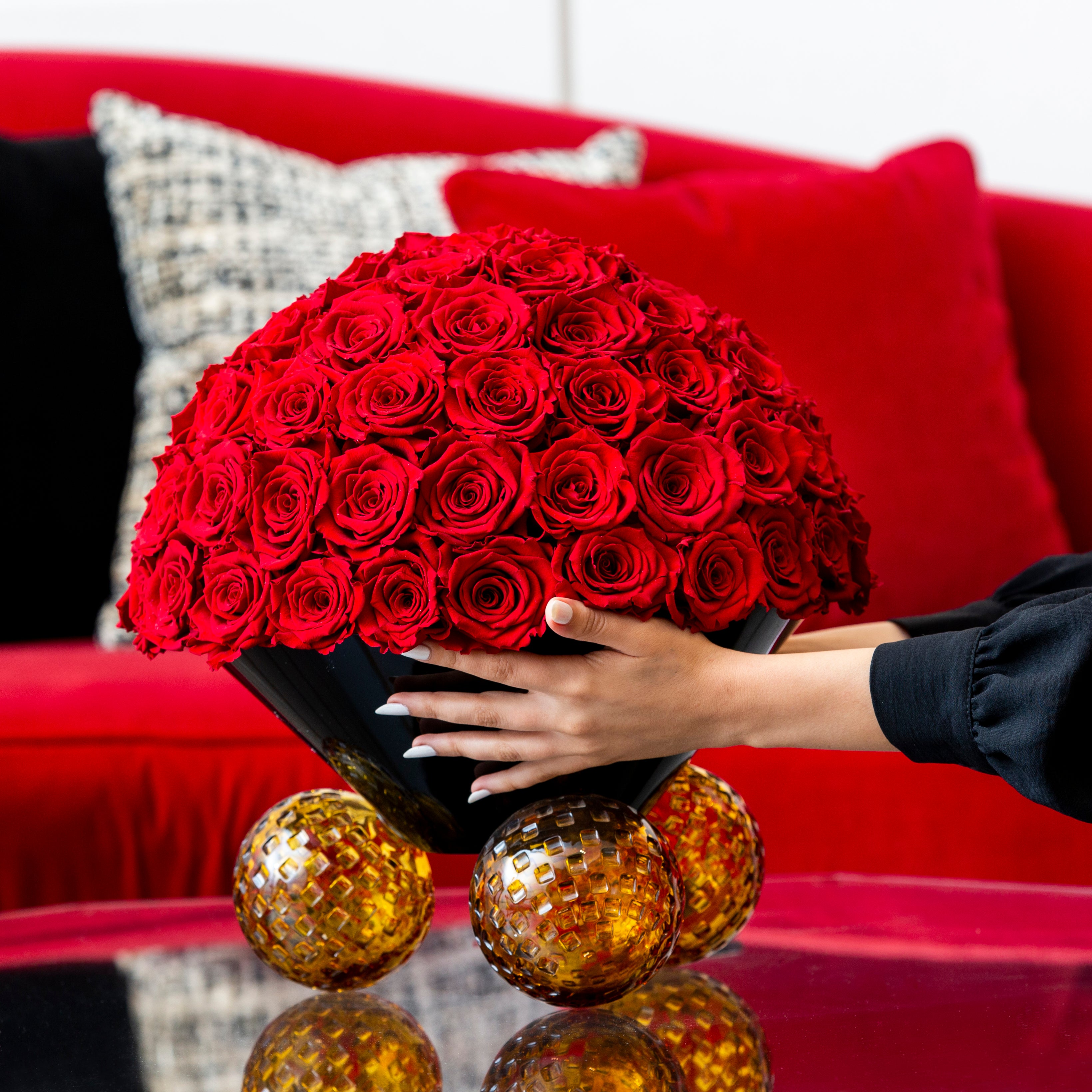 Large bouquet of red roses held by a person in front of a red couch with decorative pillows.