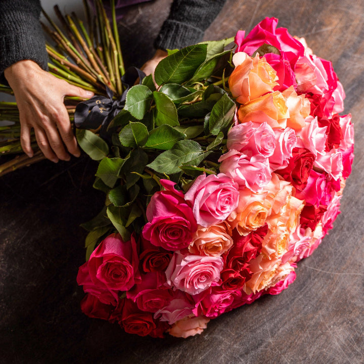 A large bouquet of fresh roses in a gradient of colors, transitioning from deep pink and red at the top to light pink and peach in the middle, with green leaves and stems, held by hands on a dark wooden surface.
