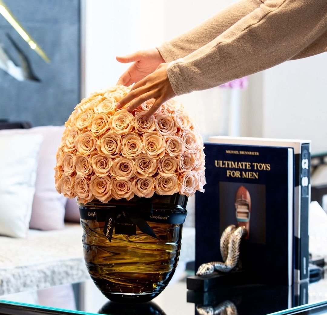 Hand touching a decorative vase filled with peach roses on a table next to a book titled 'Ultimate Toys for Men'.